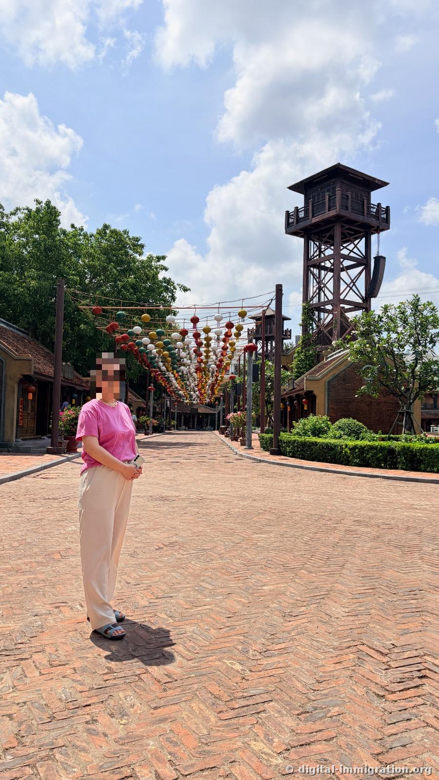 A person standing on a red brick path with colorful ball decorations hanging overhead, a tall wooden watchtower visible in the background
