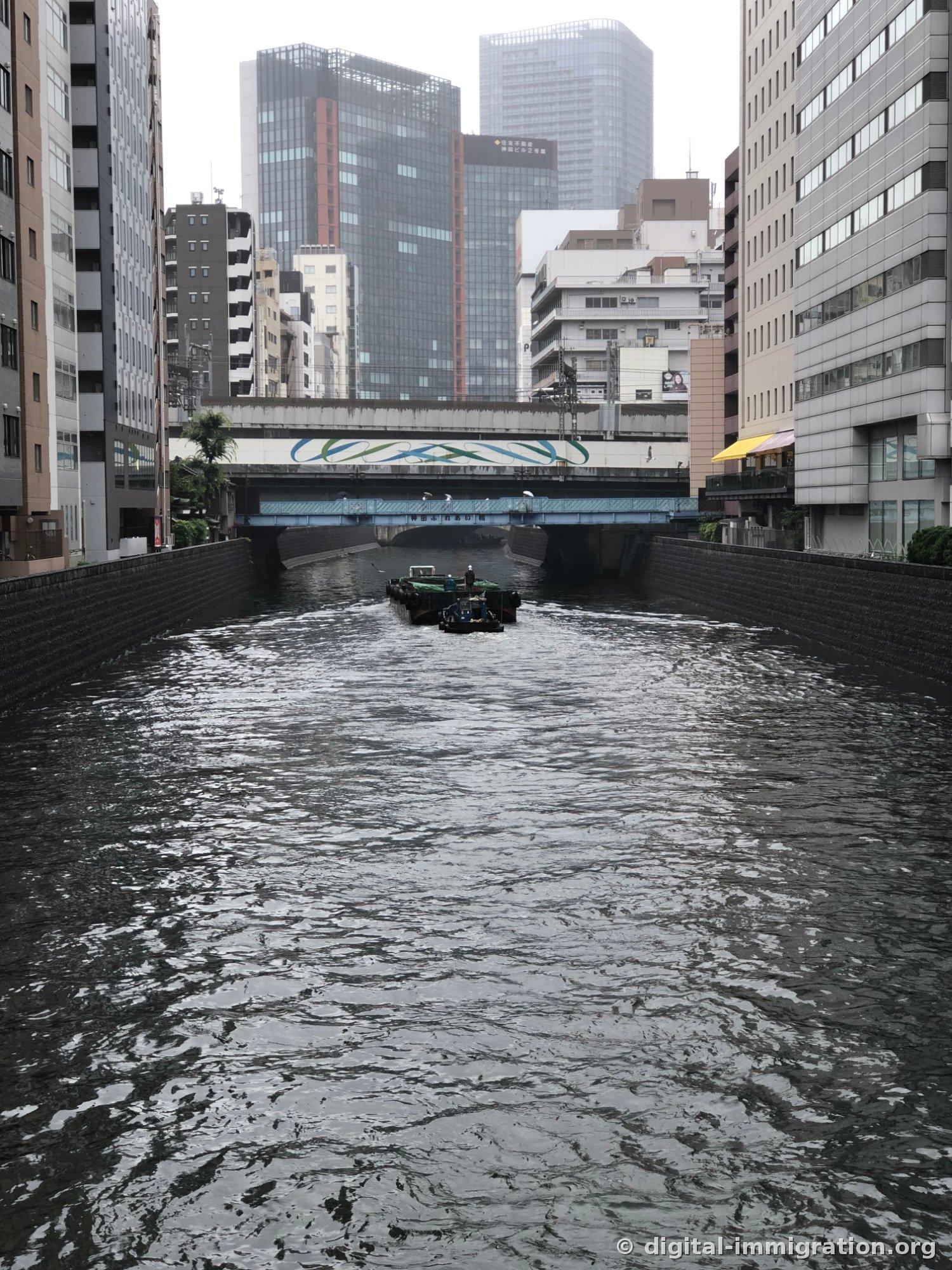 Kanda River waterway between buildings on a cloudy day with tugboat and barge passing under the bridge