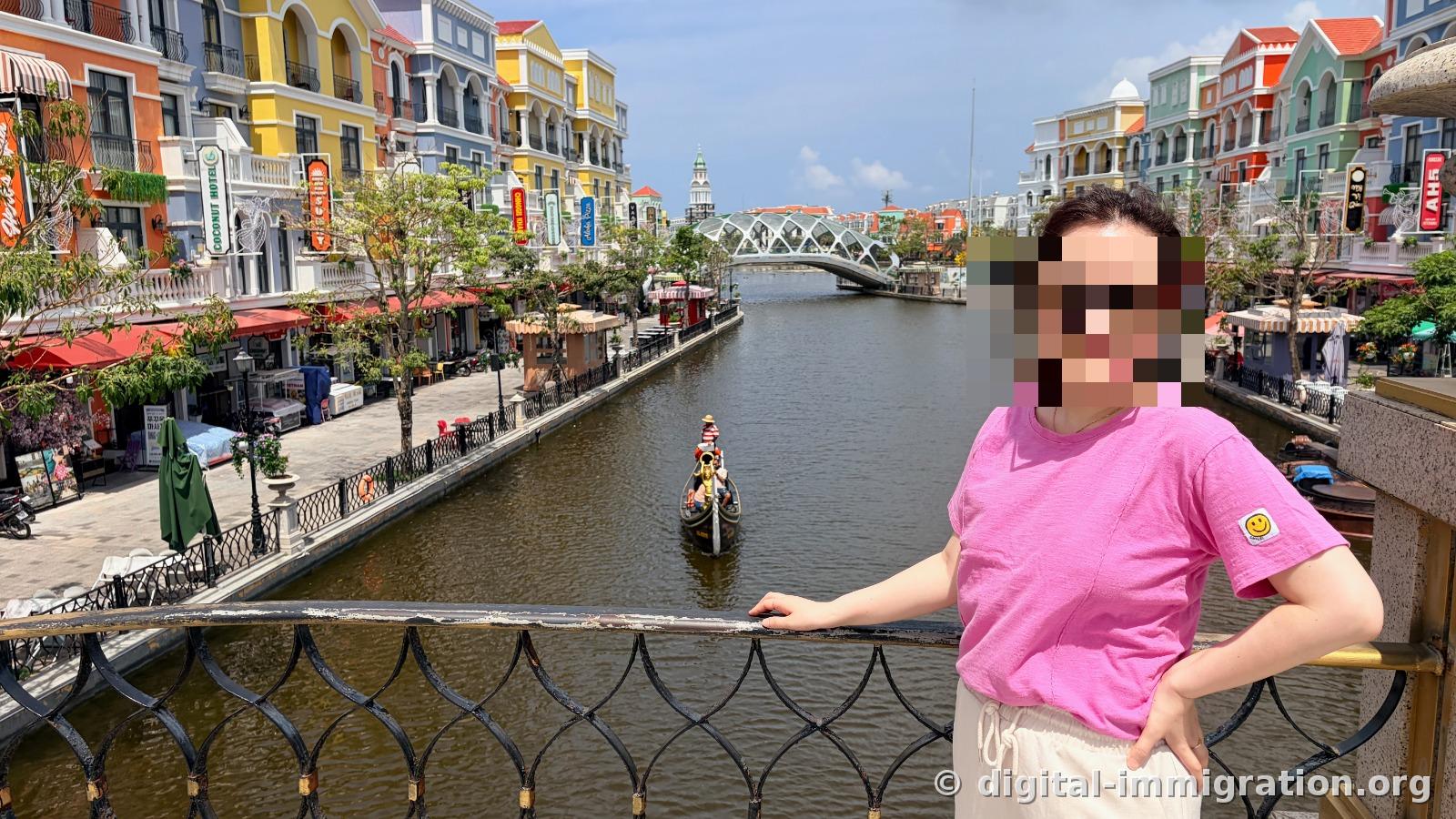 A gondola with a boatman passing under the canal, a COCONUT HOTEL sign visible on the left shop wall, a white metal arch bridge in the distance
